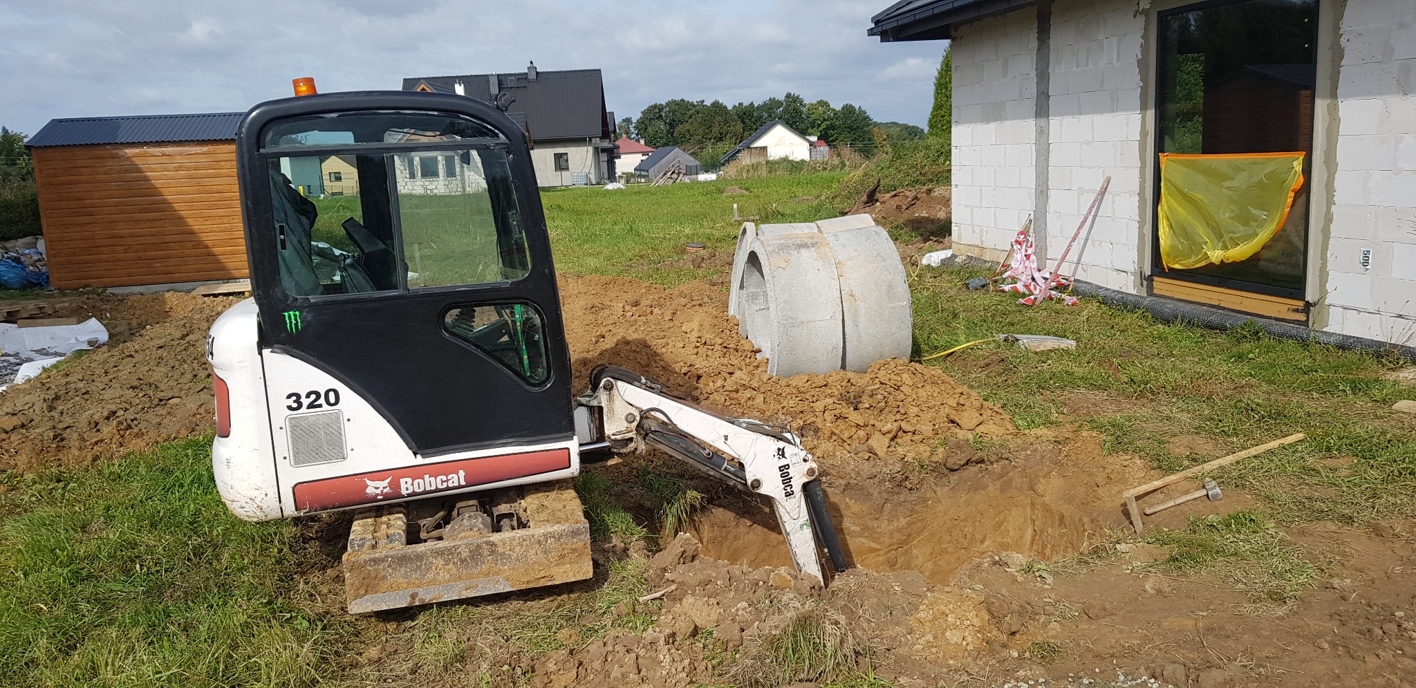 Mini koparka Bobcat 320 w trakcie wykonywania wykopu w ziemi, obok leżą betonowe kręgi, w tle budynek w trakcie budowy.