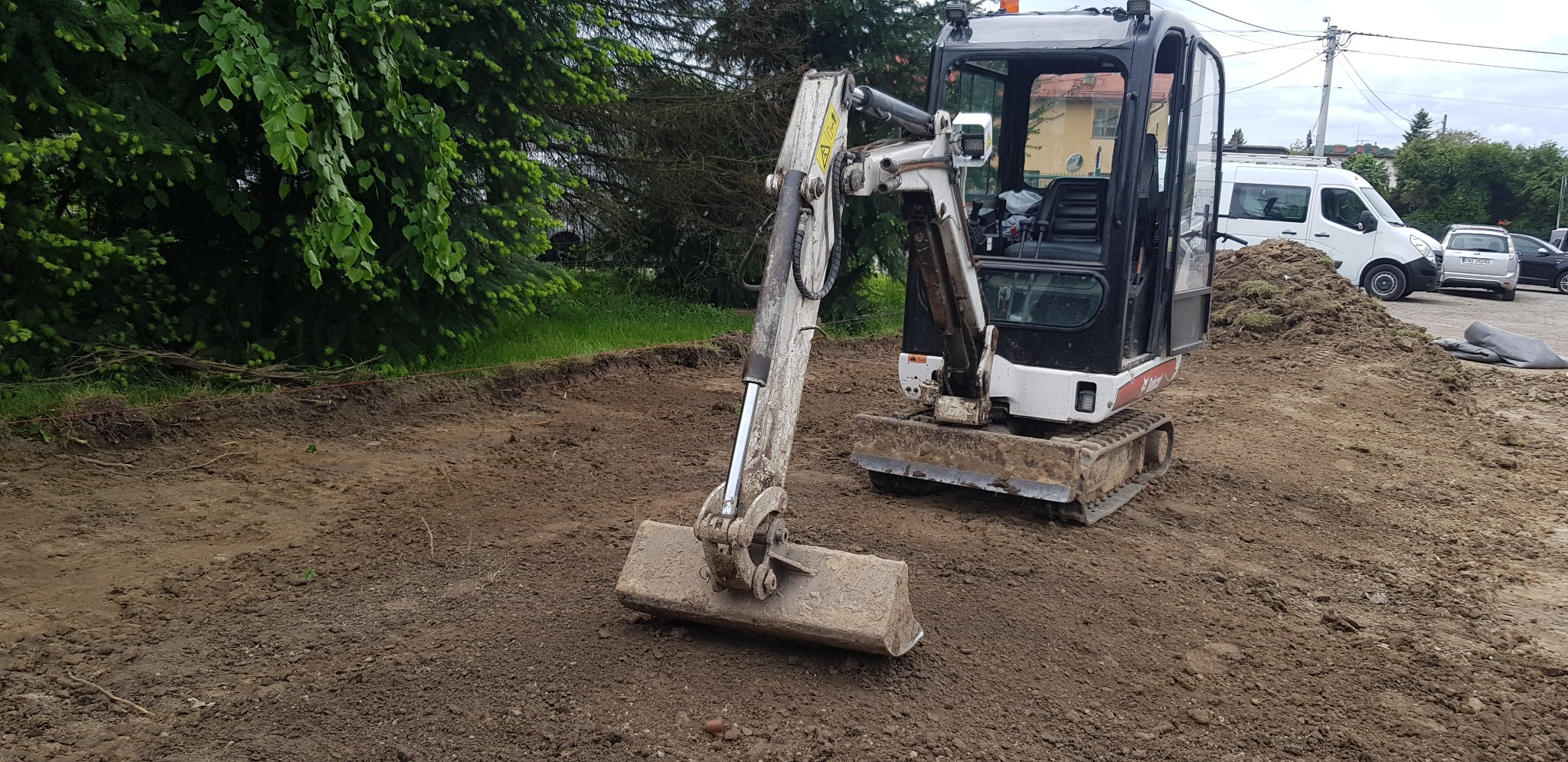 Mini koparka Bobcat na wyrównanym terenie z widoczną linią wykopu, drzewa w tle.