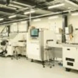 Wnętrze hali produkcyjnej z zaawansowanymi, białymi maszynami do automatycznego montażu elektroniki, widoczne czarne słupy konstrukcyjne i system wentylacji pod sufitem.