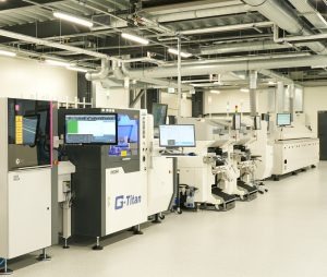 Linia produkcyjna z zaawansowanym oprzyrządowaniem, w tym maszyny z ekranami wyświetlającymi dane i interfejsy użytkownika, widoczne rury i instalacje pod sufitem, w jasnym, sterylnym pomieszczeniu.