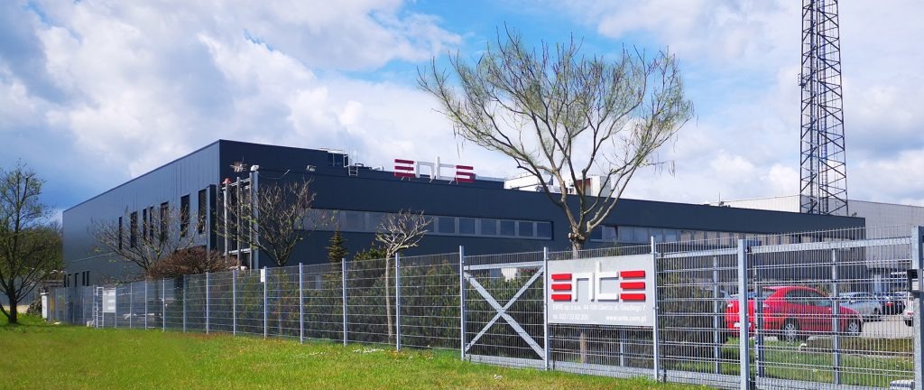 Budynek firmy IT z logo na dachu, widoczny zza ogrodzenia z tablicą informacyjną i drzewami, w tle maszt telekomunikacyjny.