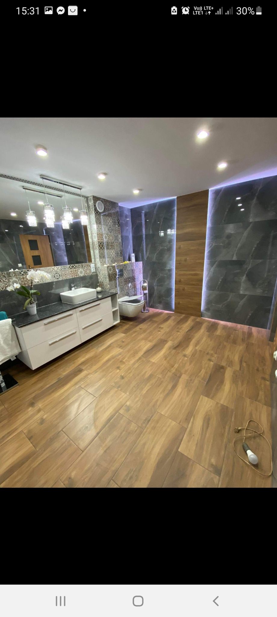 Nowoczesna łazienka z drewnianą podłogą, marmurowymi płytkami ściennymi z podświetleniem LED, białą umywalką na szafce z szufladami i lustrem z wiszącymi lampami.