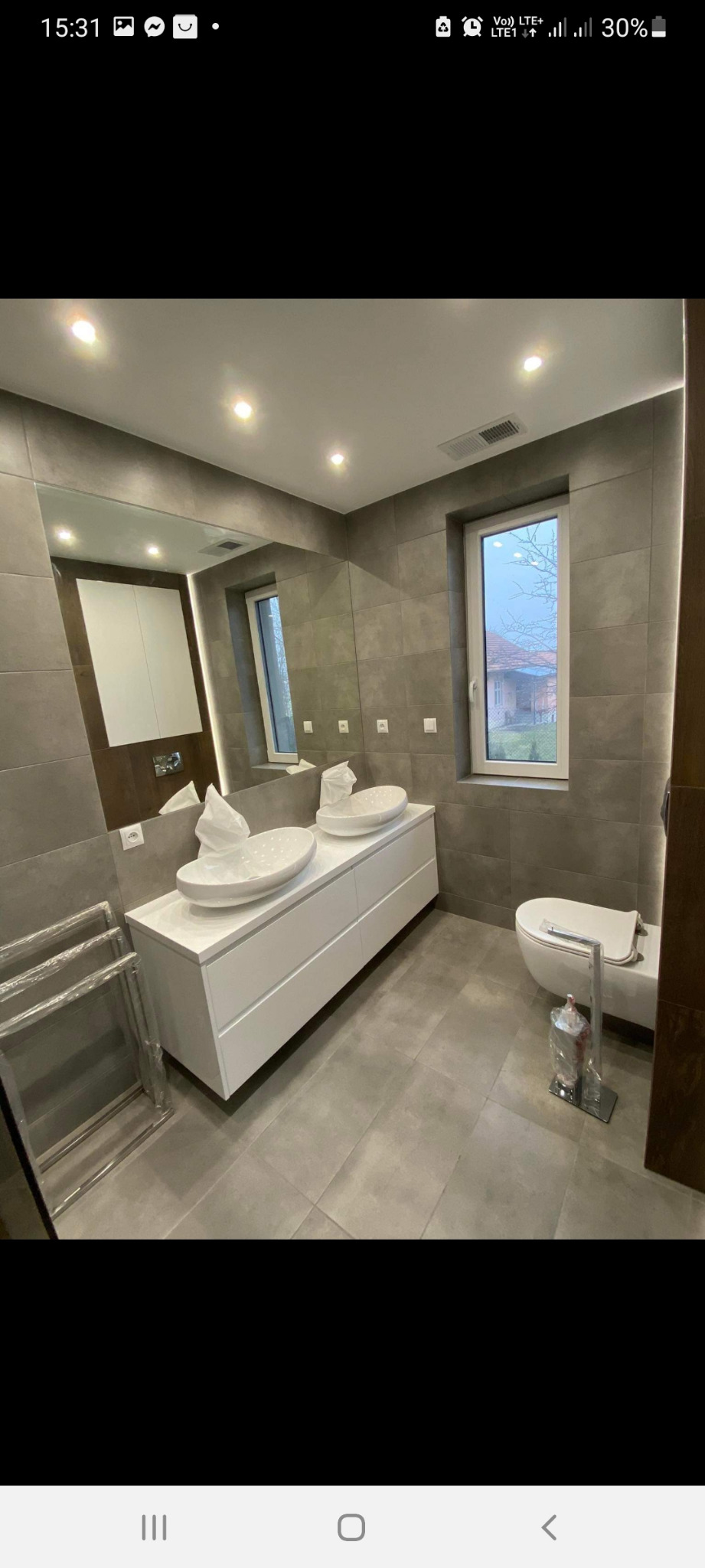 Wykończona łazienka w szarościach z podwójną umywalką nablatową, dużym lustrem, oświetleniem punktowym i podświetleniem LED oraz nową toaletą. Na podłodze i ścianach duże, kwadratowe płytki. W tle...