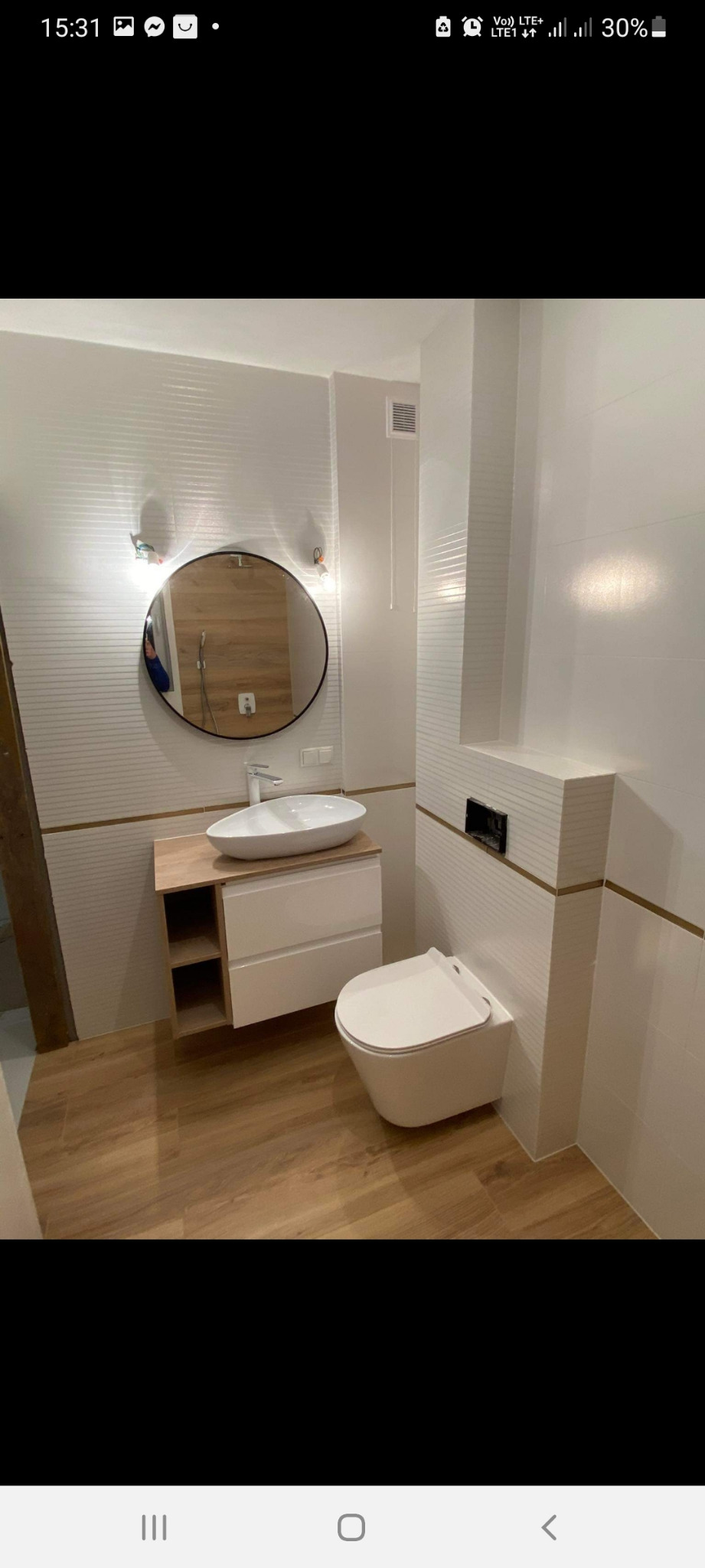 Nowoczesna łazienka z wiszącą toaletą, umywalką nablatową na szafce z drewnianym blatem i okrągłym lustrem podświetlanym kinkietami, wykończona płytkami z poziomymi paskami.