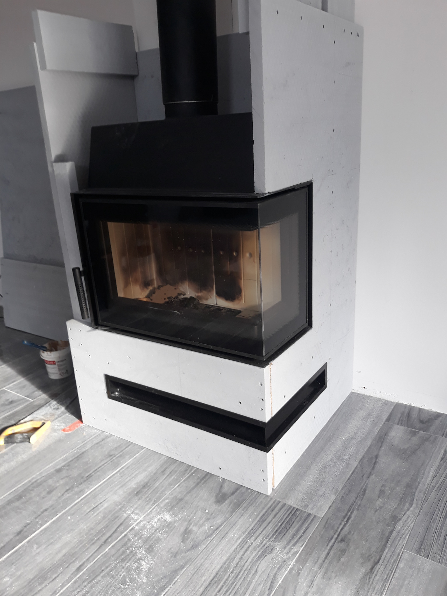 Wnętrze z kominkiem obudowanym białymi płytami, widoczne narzędzia i resztki kleju na podłodze z szarych paneli imitujących drewno, sugerujące prace wykończeniowe.