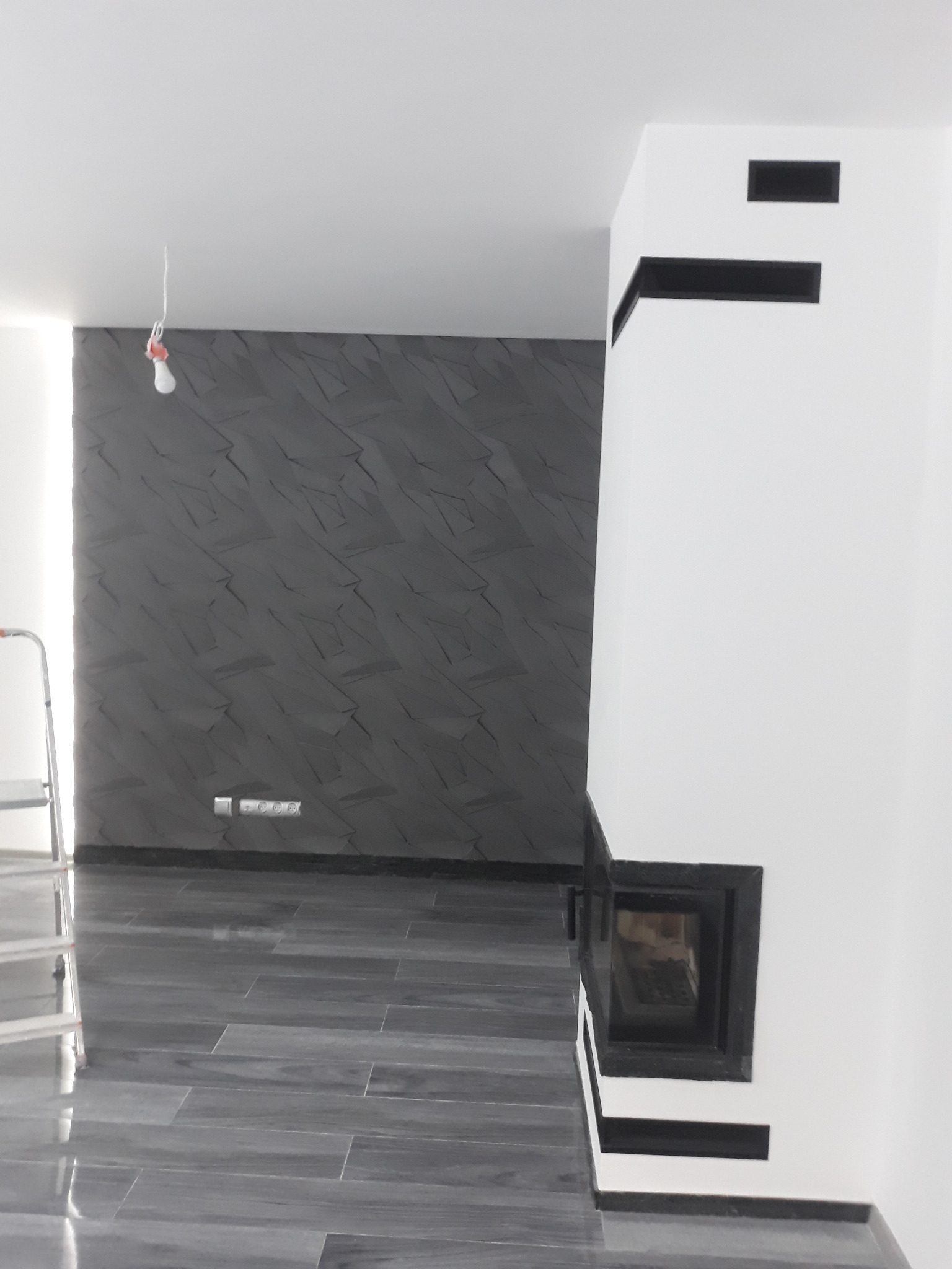 Wnętrze pokoju w Nowym Sączu z szarą ścianą o geometrycznym wzorze, ciemną podłogą z płytek imitujących drewno, białym kominkiem z czarnymi akcentami i wiszącą żarówką.