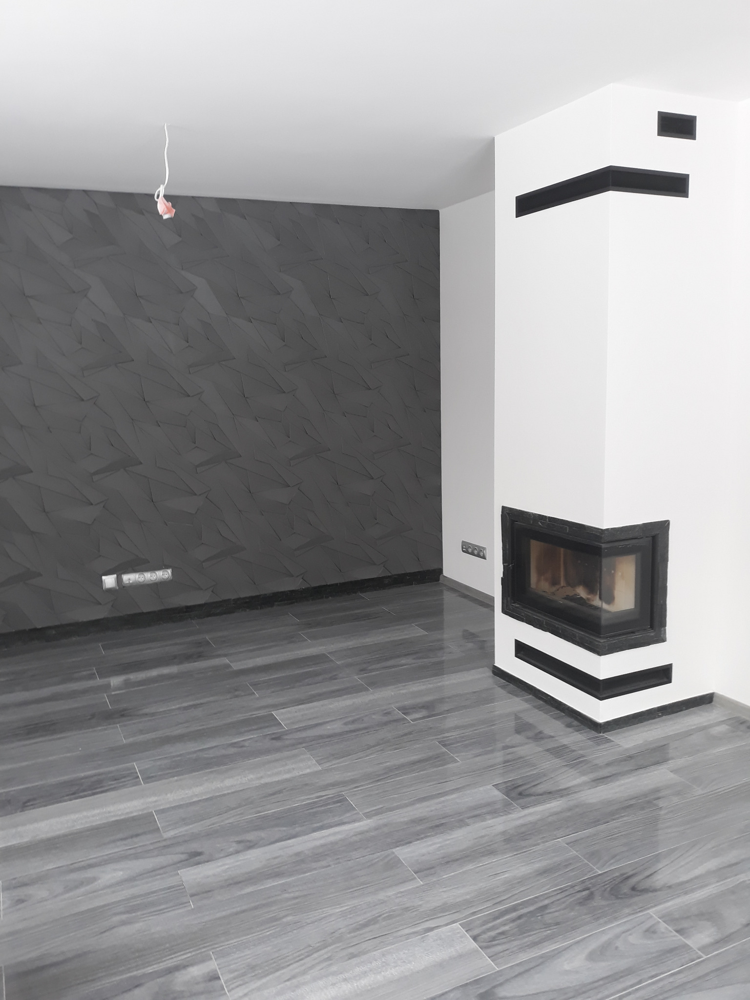 Szare płytki podłogowe imitujące drewno w nowocześnie wykończonym salonie z kominkiem, ściana z trójwymiarową strukturą, widoczne gniazdka elektryczne i niedokończone oświetlenie sufitowe.