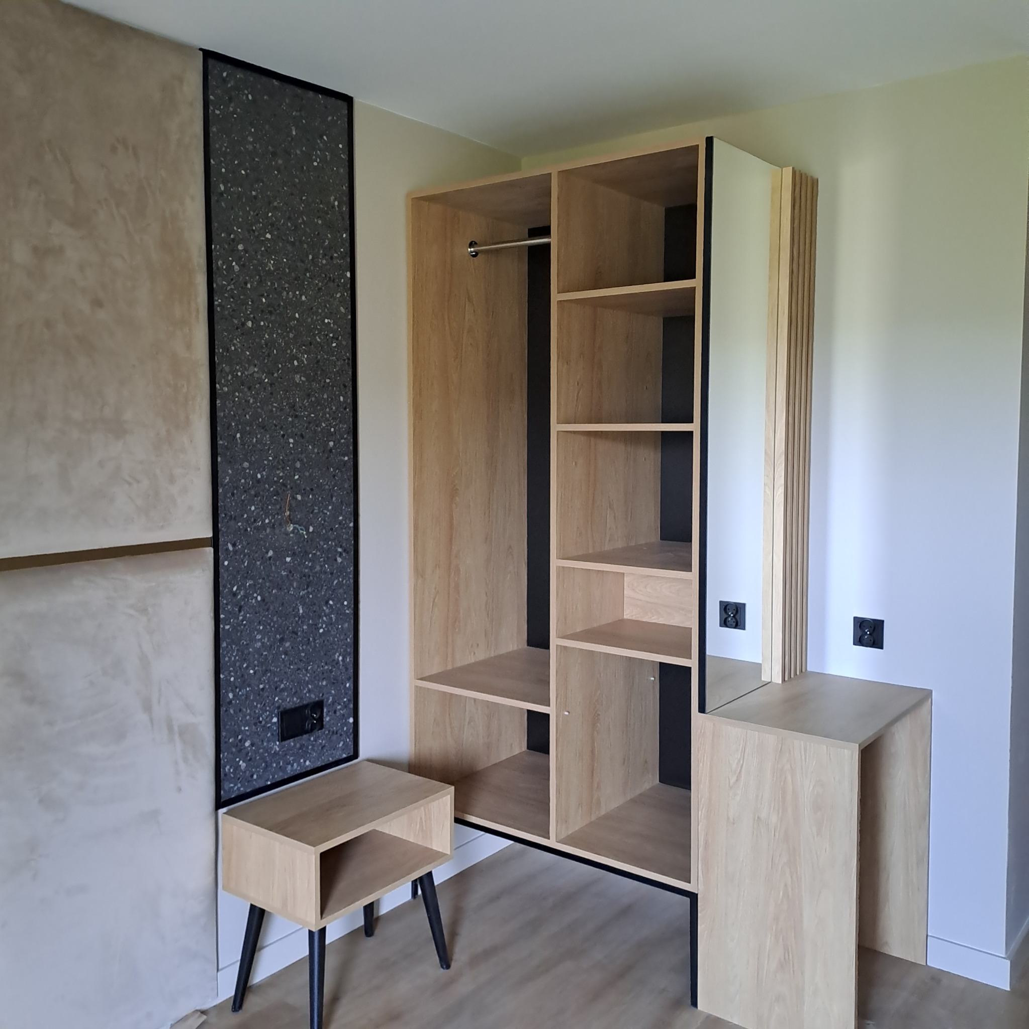 Drewniana szafa z półkami i drążkiem, stolik nocny na czarnych nóżkach oraz drugi stolik nocny w jasnym pokoju z beżową ścianą i panelem dekoracyjnym.