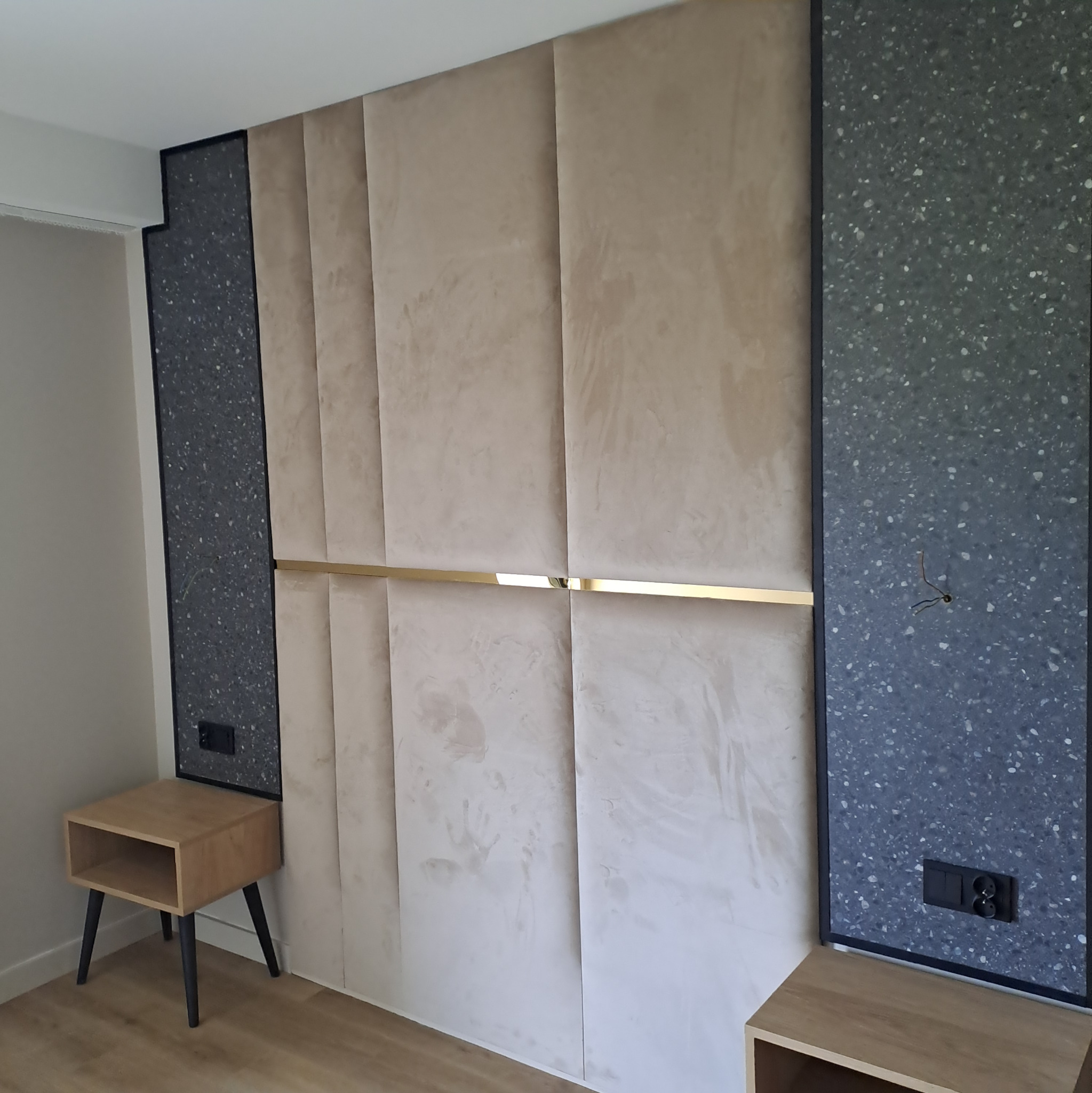 Beżowe panele tapicerowane na ścianie z poziomym, złotym akcentem, otoczone panelami z szarego lastryko. Widoczne gniazdka elektryczne i drewniane szafki nocne.