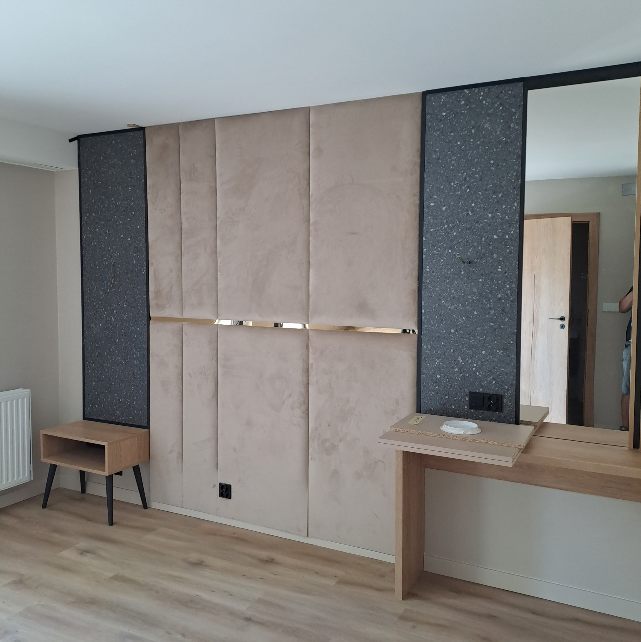 Beżowe panele ścienne z złotymi akcentami i szarymi panelami bocznymi w nowoczesnym wnętrzu, obok drewniana szafka nocna i toaletka z lustrem.