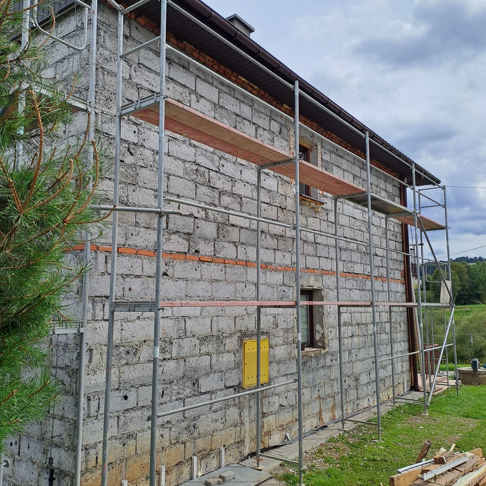Budynek z szarego bloczka z widocznymi śladami po starym murze z czerwonej cegły, otoczony metalowym rusztowaniem, w tle zieleń i pochmurne niebo.