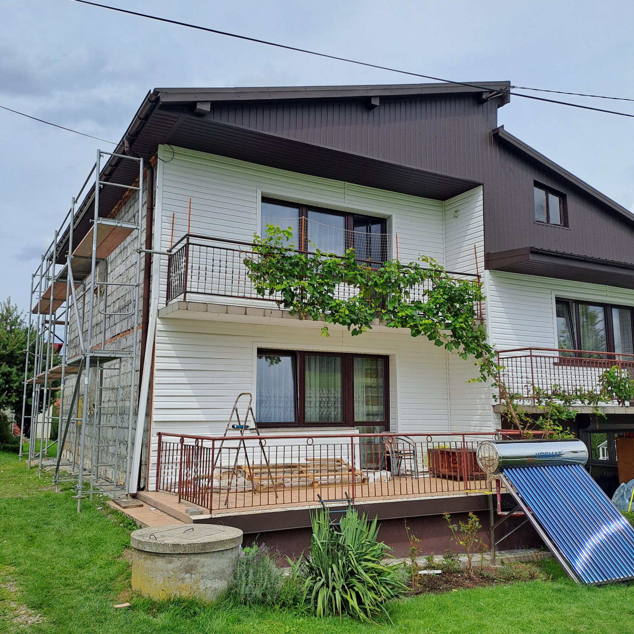 Dwupiętrowy dom z białą elewacją sidingową w trakcie remontu, z rusztowaniem przy ścianie szczytowej, balkonem porośniętym winoroślą i kolektorem słonecznym na trawniku.