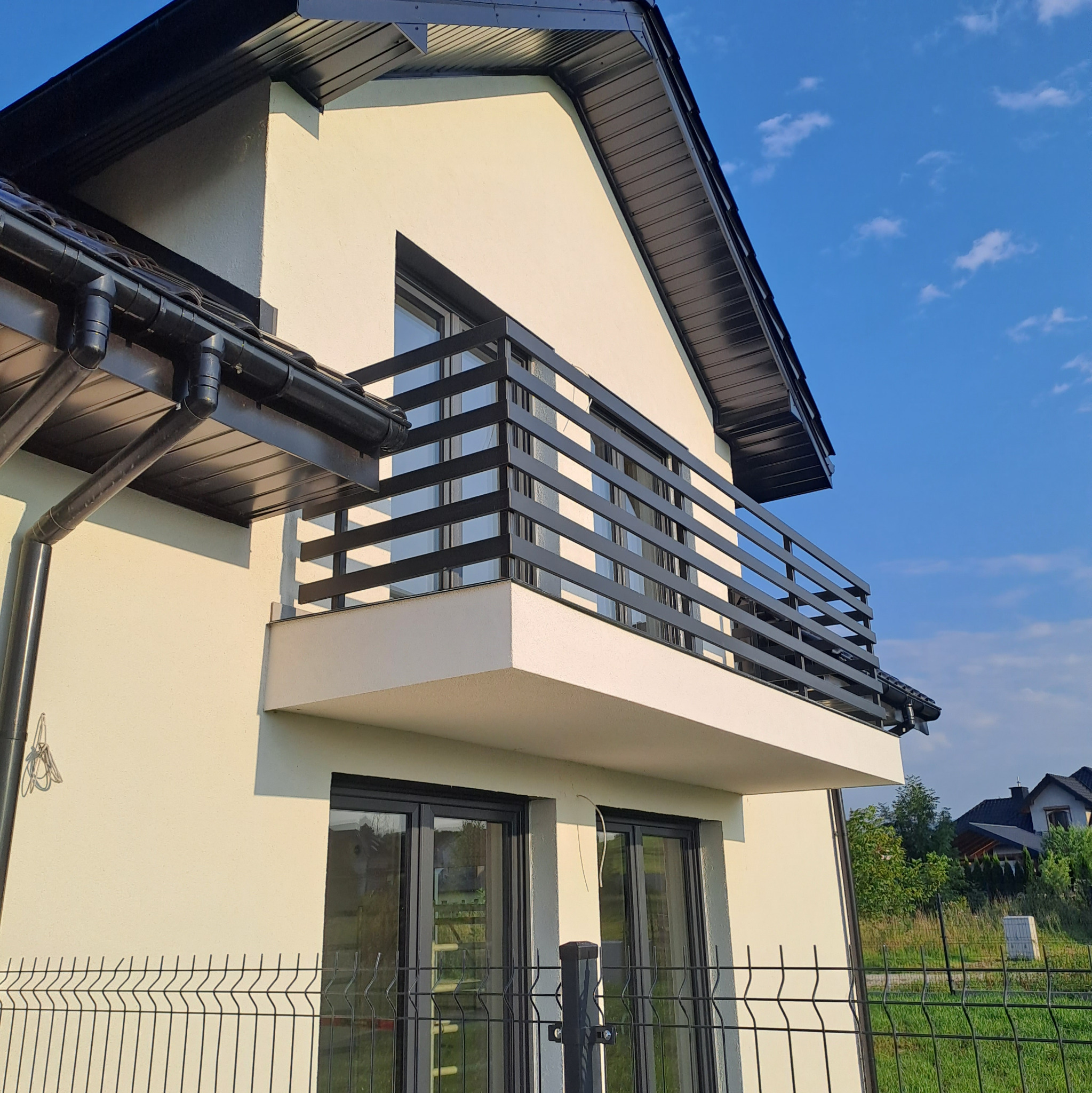 Nowoczesny dom jednorodzinny z balkonem z czarną balustradą, kremowa elewacja, czarne rynny i okna, widok z dołu na tle błękitnego nieba.