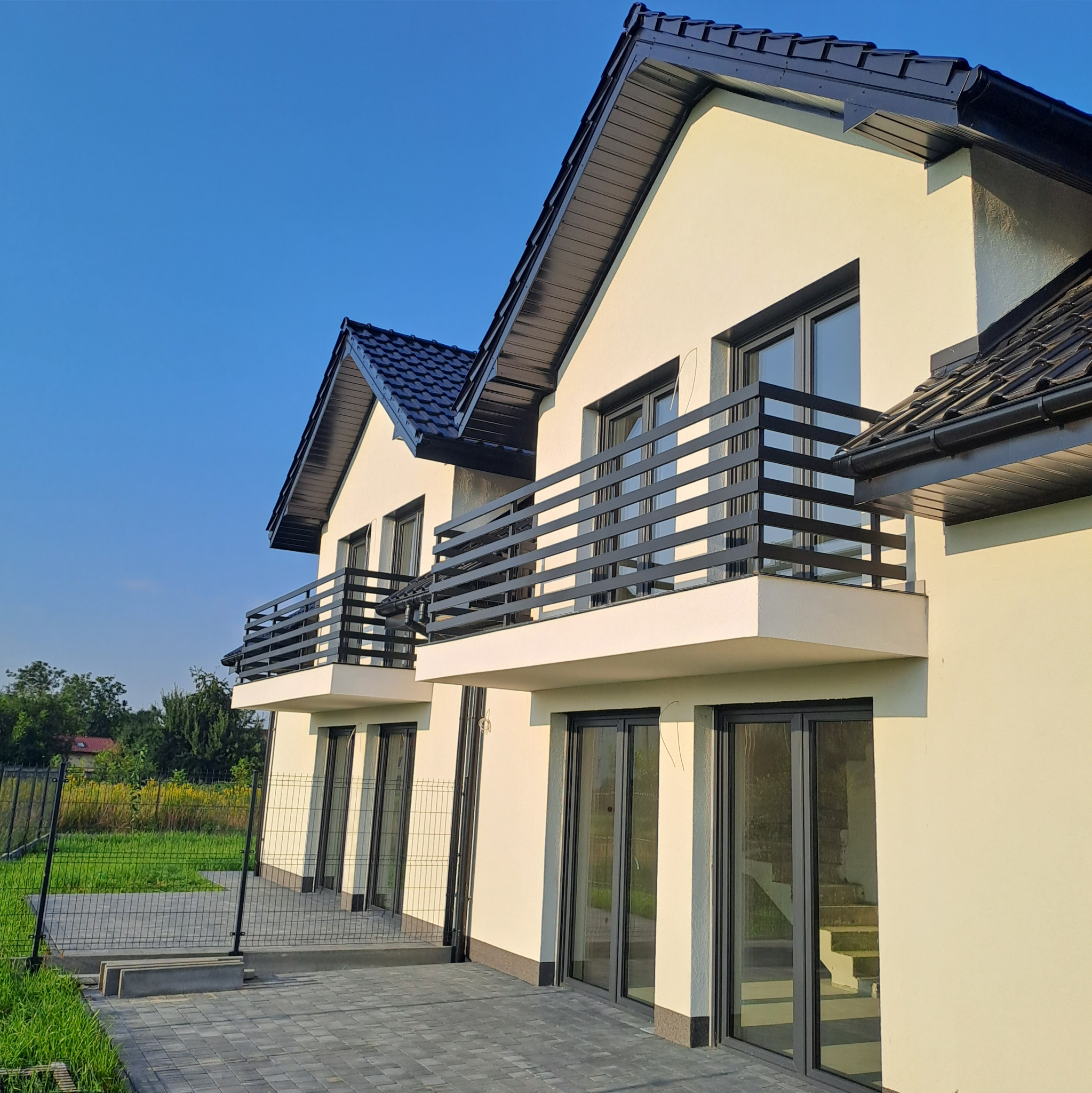 Nowoczesny dom jednorodzinny z jasną elewacją, czarnymi ramami okiennymi i balkonami z metalowymi balustradami, widok z zewnątrz na tle błękitnego nieba.