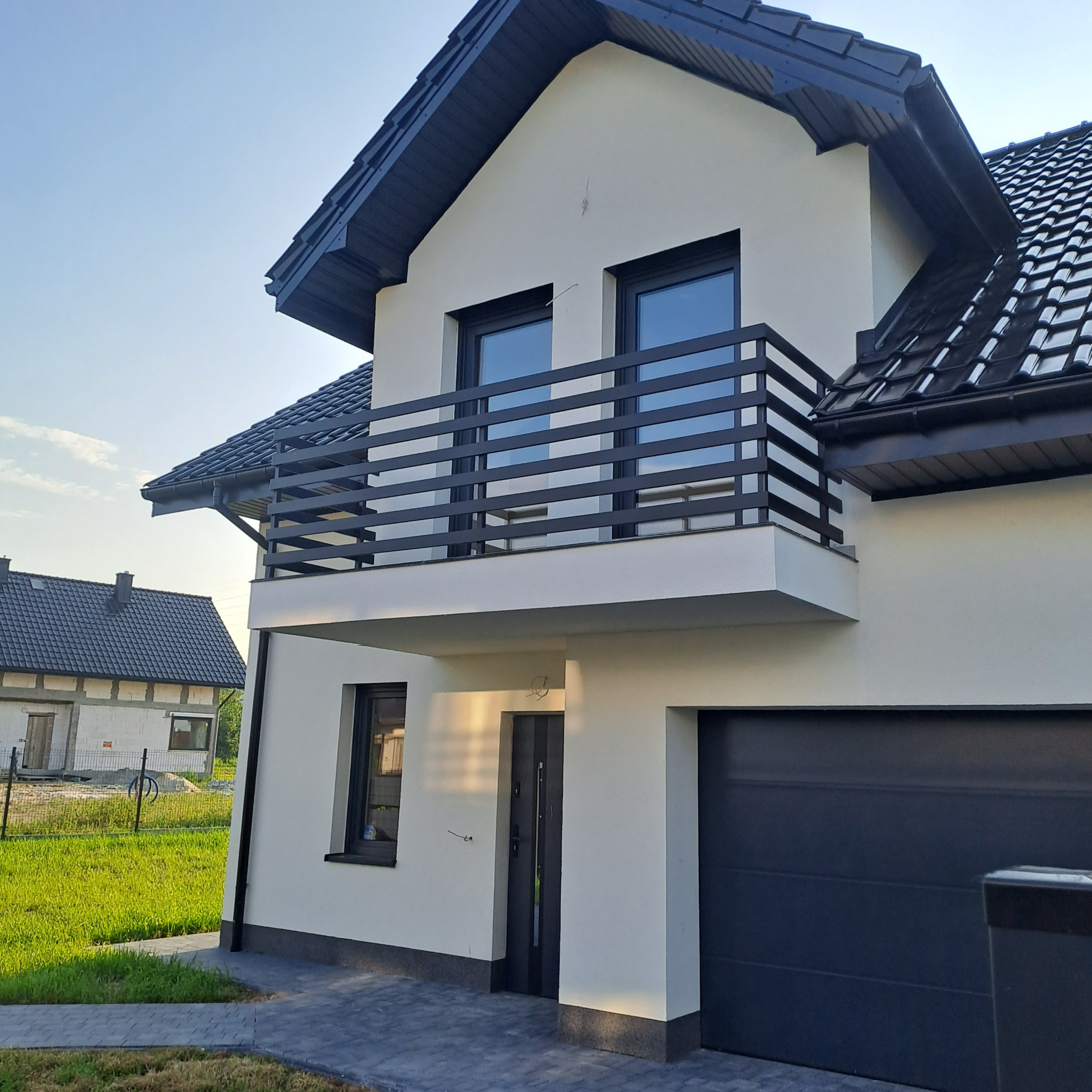 Nowoczesny dom jednorodzinny w Nowym Sączu z balkonem z czarną balustradą, białą elewacją i ciemną stolarką okienną oraz automatyczną bramą garażową.