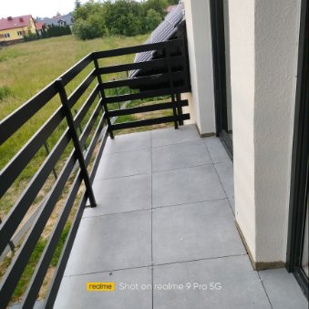Nowoczesny balkon wykończony szarymi płytkami ceramicznymi, z czarną metalową balustradą i widokiem na zieleń w tle.