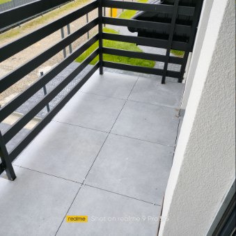 Szary balkon wyłożony kwadratowymi płytkami z ciemną fugą, czarna metalowa balustrada, widok z góry, w tle trawnik.