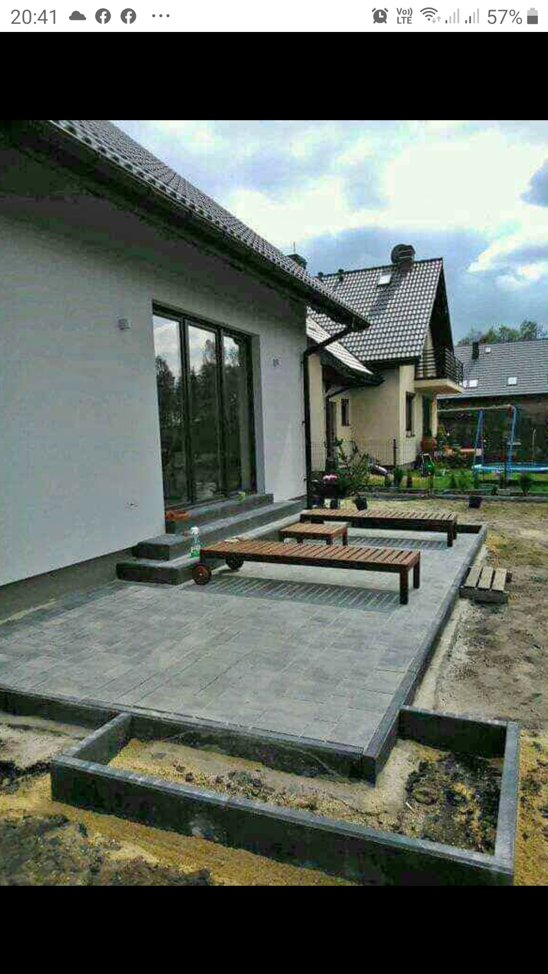 Ułożona szara kostka brukowa na tarasie przed domem jednorodzinnym, widoczne schody wejściowe, drewniane leżaki na kółkach oraz przygotowane obrzeże pod rabatę kwiatową.