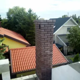 Wzniesiony z cegły klinkierowej komin na tle budynków mieszkalnych z dachami krytymi czerwoną dachówką oraz fragmentu ściany z białego betonu komórkowego.