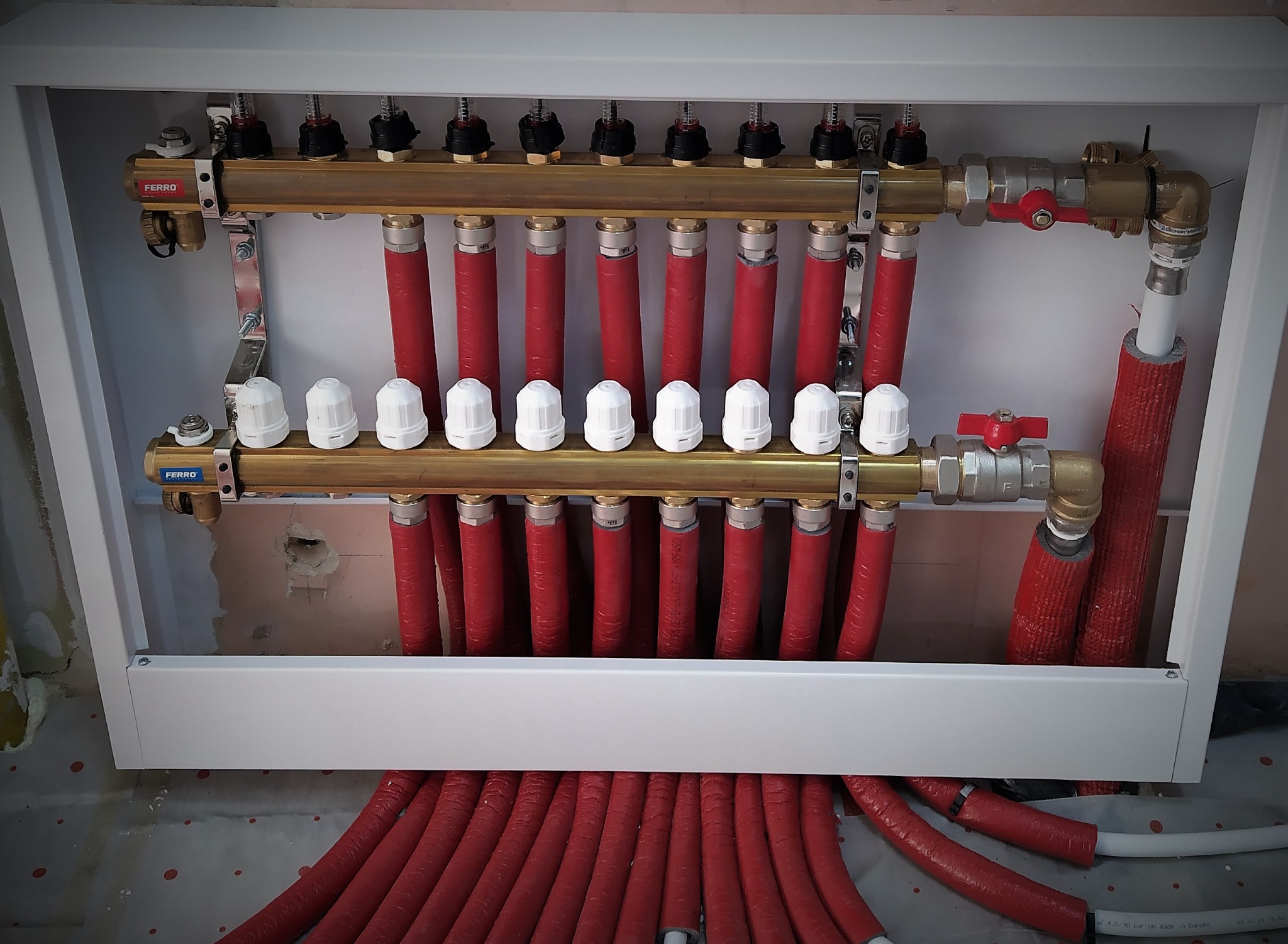 Rozdzielacz ogrzewania podłogowego w białej skrzynce, widoczne czerwone rury grzewcze z izolacją i mosiężne elementy łączeniowe marki Ferro.
