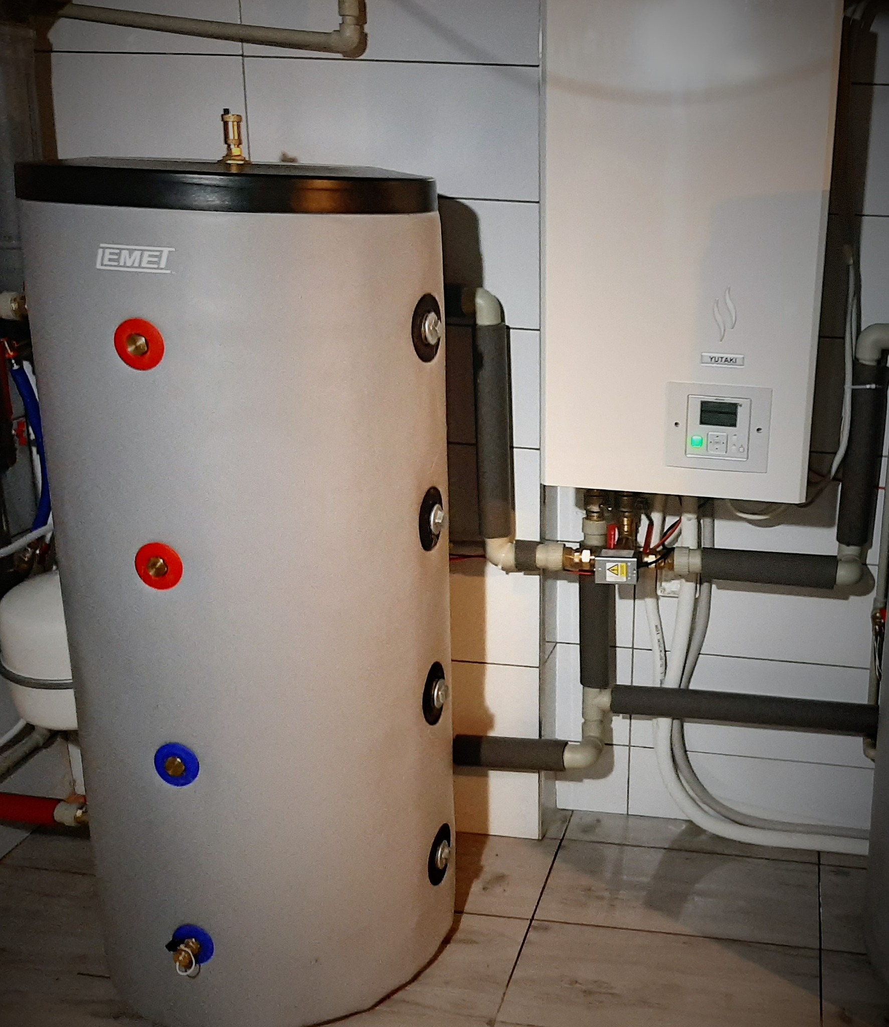 Szary zbiornik akumulacyjny ciepła marki Emet z widocznymi przyłączami oznaczonymi kolorami, obok biały piec grzewczy Yutaki z wyświetlaczem i podłączeniami rur w pomieszczeniu z wykafelkowanymi...