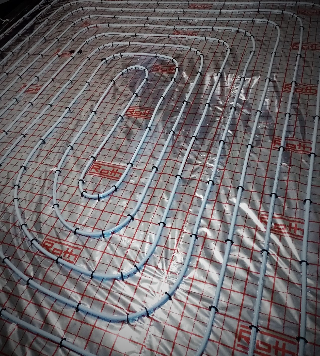 Białe rurki ogrzewania podłogowego Roth, zamocowane czarnymi klipsami do folii z czerwoną siatką, ułożone w pętle na dużej powierzchni.