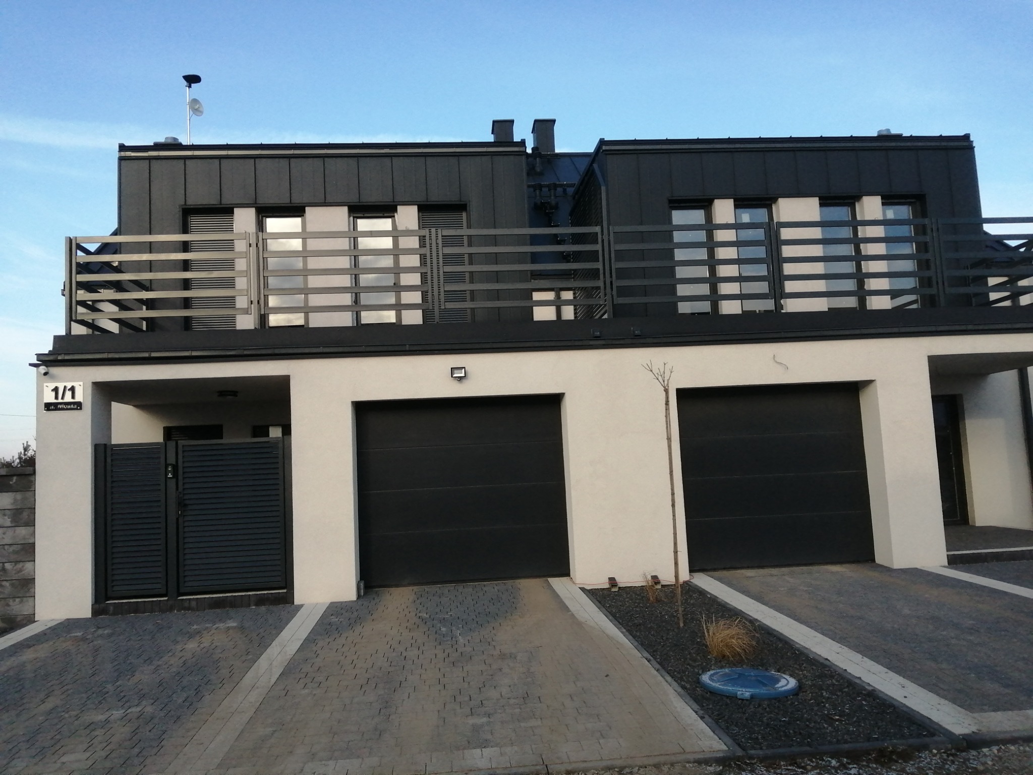 Nowoczesny dom bliźniak z czarnymi bramami garażowymi, grafitową elewacją na piętrze, balkonem z metalowymi barierkami i podjazdem wyłożonym szarą kostką brukową.