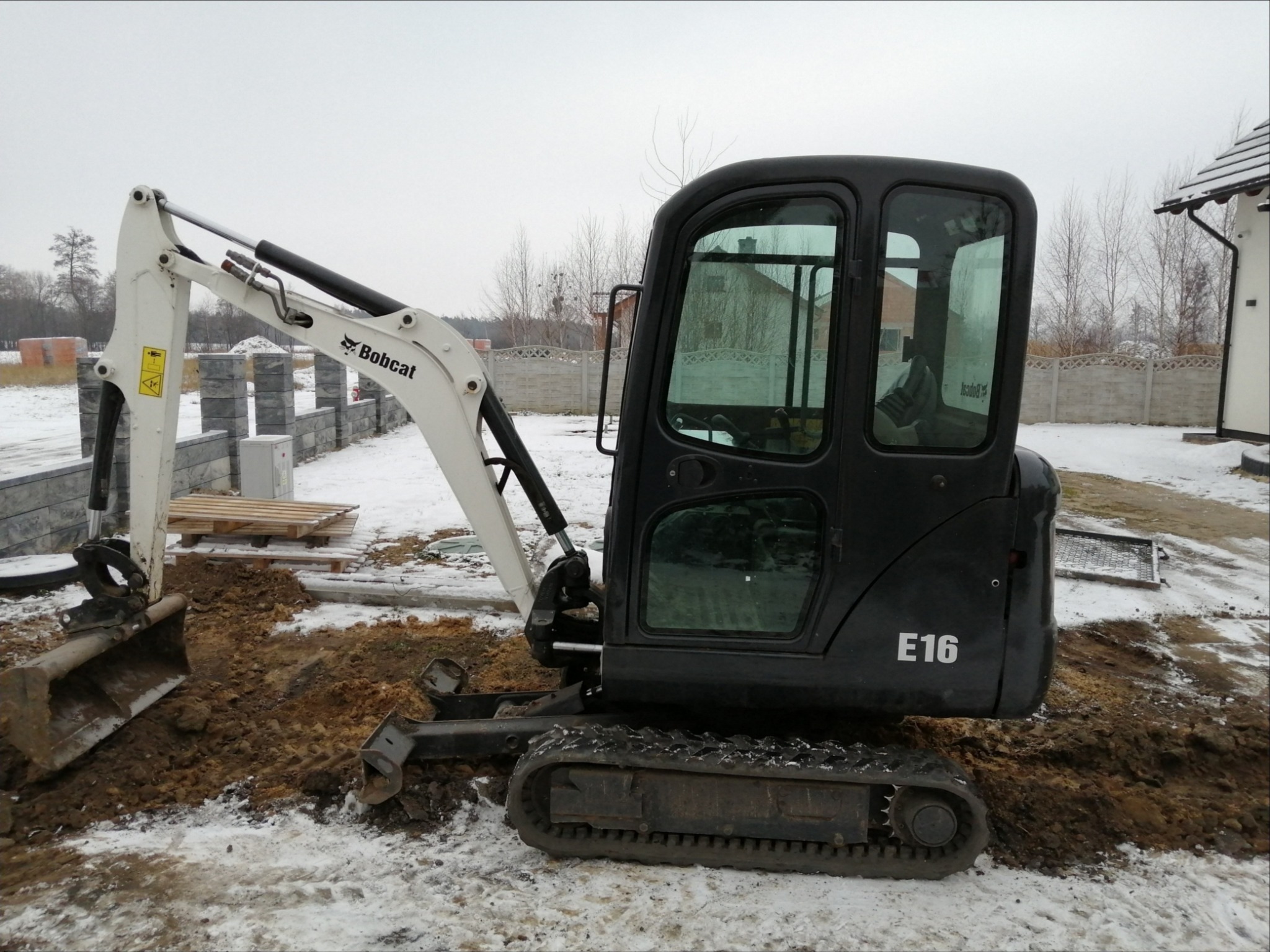 Czarny mini-koparka Bobcat E16 na placu budowy w zimowej scenerii, obok sterty kostki brukowej i palety, widoczny wykop w ziemi pokrytej śniegiem.