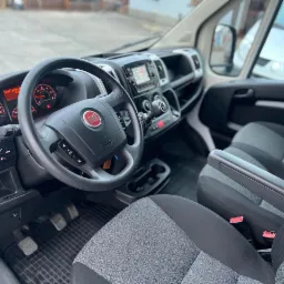 Wnętrze dostawczego Fiata Ducato z szaro-czarną tapicerką, widok z perspektywy pasażera na deskę rozdzielczą, kierownicę z logo Fiat i częściowo widoczne fotele.