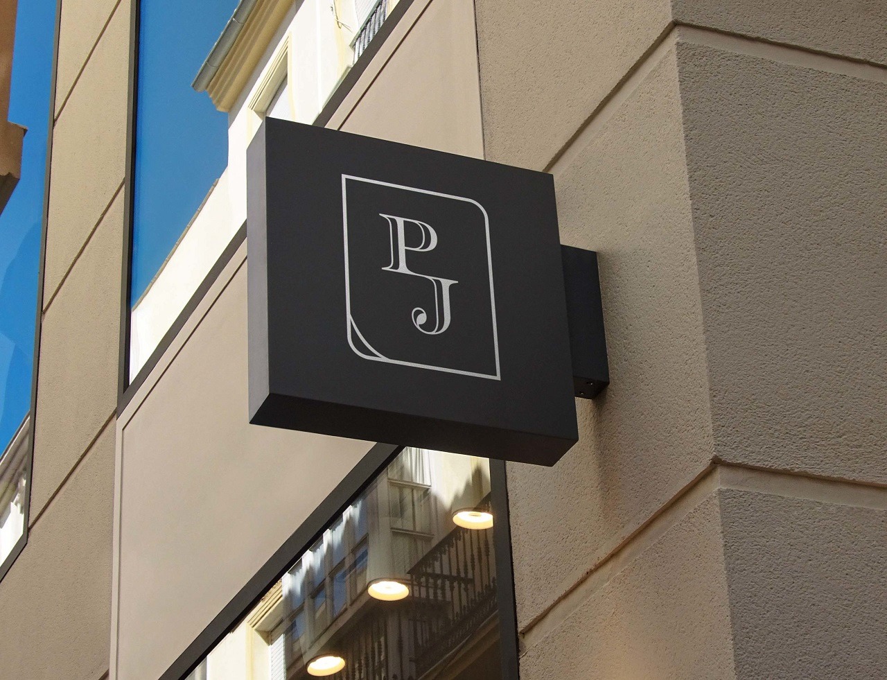 Czarny szyld reklamowy z białym logo PJ na fasadzie budynku, widok z dołu.