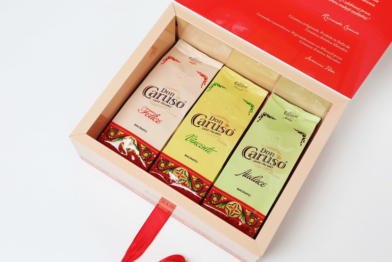 Eleganckie opakowanie prezentowe z trzema paczkami mielonej kawy Don Caruso w kremowych, żółtych i zielonych torebkach, każda z innym smakiem, ozdobione czerwoną wstążką i wzorem ceramicznym.