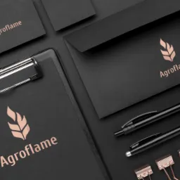 Czarny zestaw materiałów biurowych z logo Agrofame w kolorze miedzianym, zawierający wizytówkę, kopertę, notatnik z klipem, dwa długopisy i spinacze w kolorze różowego złota, ułożone na ciemnym tle.