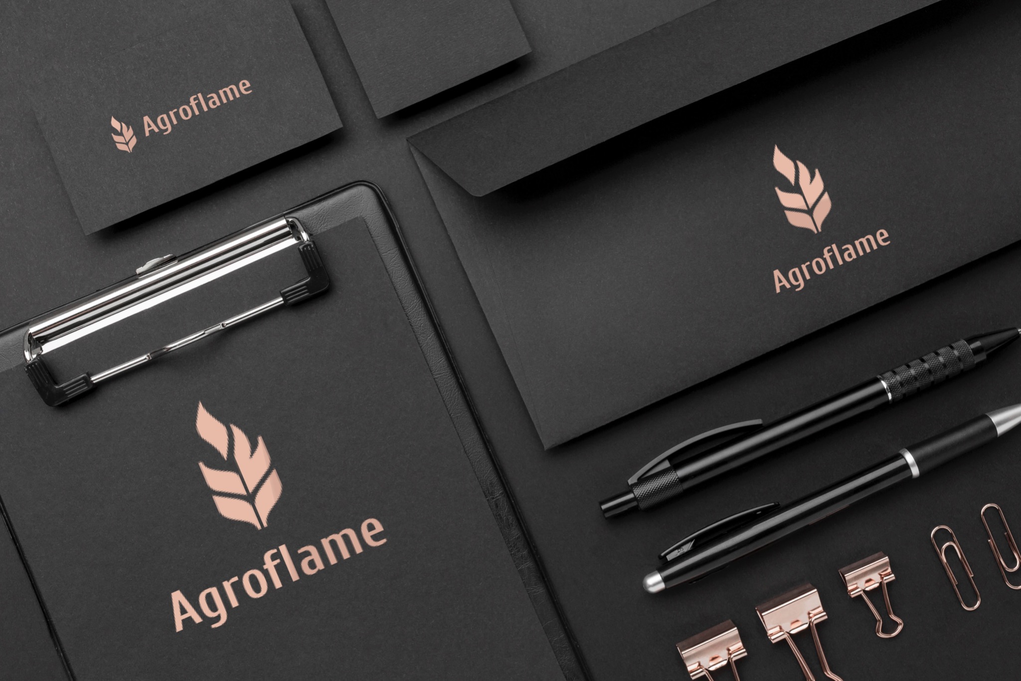 Czarny zestaw materiałów biurowych z logo Agrofame w kolorze miedzianym, zawierający wizytówkę, kopertę, notatnik z klipem, dwa długopisy i spinacze w kolorze różowego złota, ułożone na ciemnym tle.