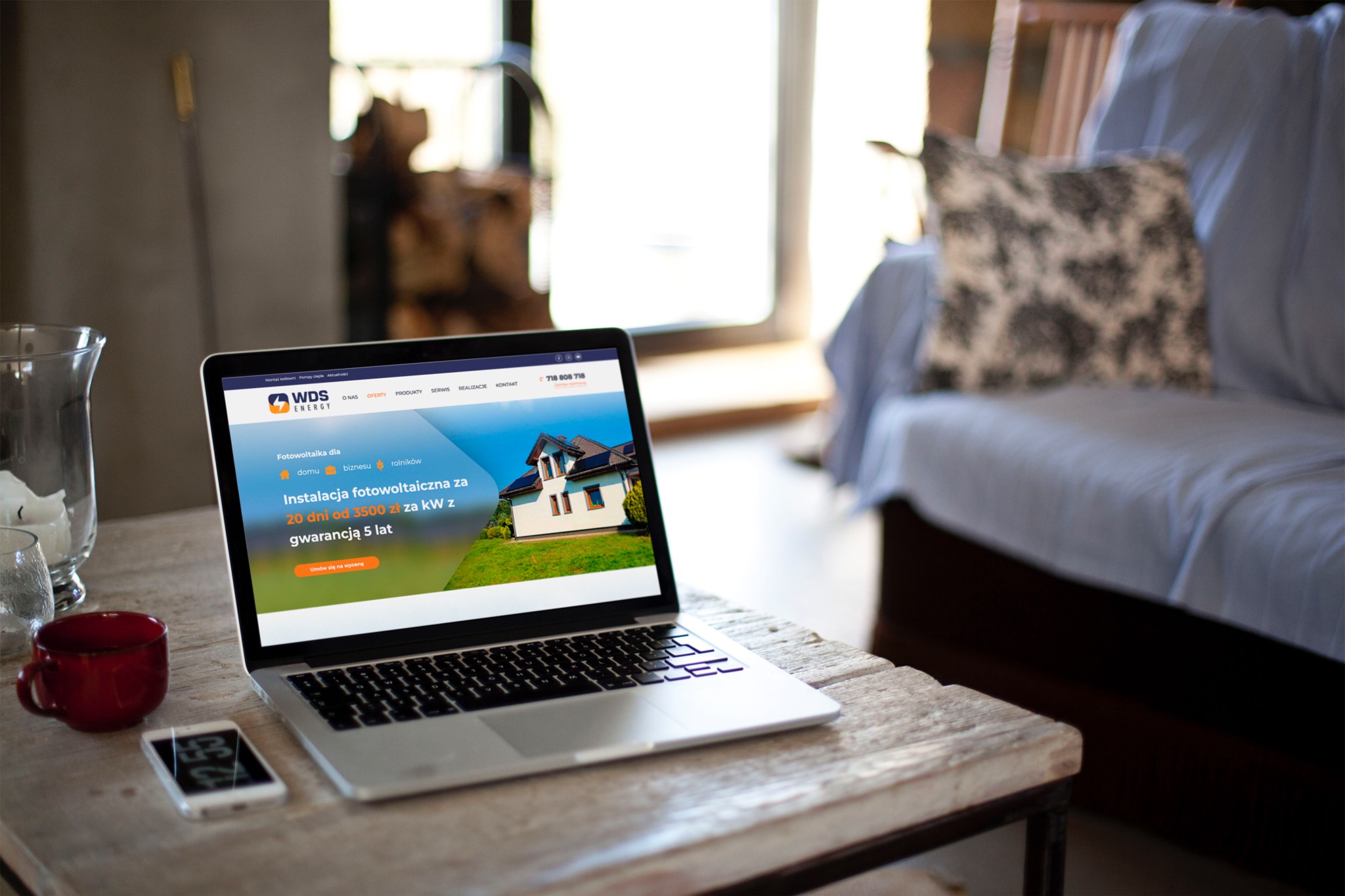 Laptop wyświetlający stronę internetową firmy WDS Energy oferującej instalacje fotowoltaiczne, obok czerwony kubek i smartfon na drewnianym stoliku, w tle rozmyte wnętrze pokoju z kanapą.