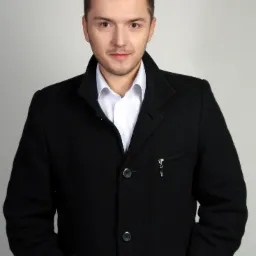 Portret młodego mężczyzny w czarnym płaszczu i białej koszuli, z rękami w kieszeniach, na jasnoszarym tle.