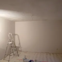 Pokój po szpachlowaniu i malowaniu, widoczne dwie drabiny, lampa halogenowa, ślady gładzi na podłodze, okno z grzejnikiem, ściany w jasnym kolorze.
