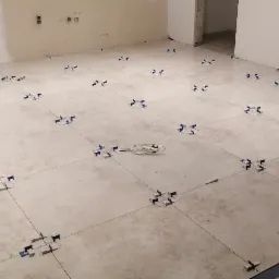 Kremowe płytki podłogowe w trakcie układania, z systemem poziomowania płytek (klipsy i kliny) rozmieszczonymi na całej powierzchni, widoczne gniazdka elektryczne w ścianie i przewód elektryczny...