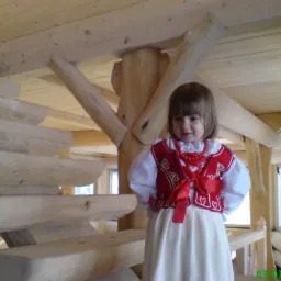 Dziewczynka w stroju ludowym stoi na drewnianych schodach wewnątrz domu z bali, widoczne elementy konstrukcyjne dachu i okno.