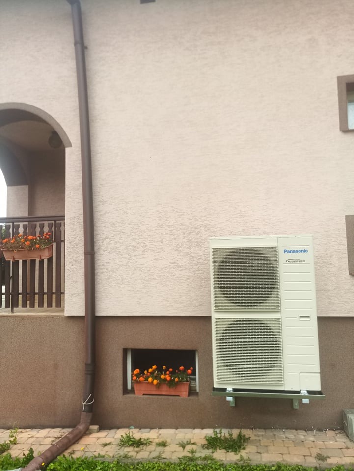 Zewnętrzna jednostka klimatyzacji Panasonic Inverter zamontowana na ścianie budynku obok okna piwnicznego z doniczką pomarańczowych kwiatów, widoczny także fragment rynny i balkon z kwiatami.