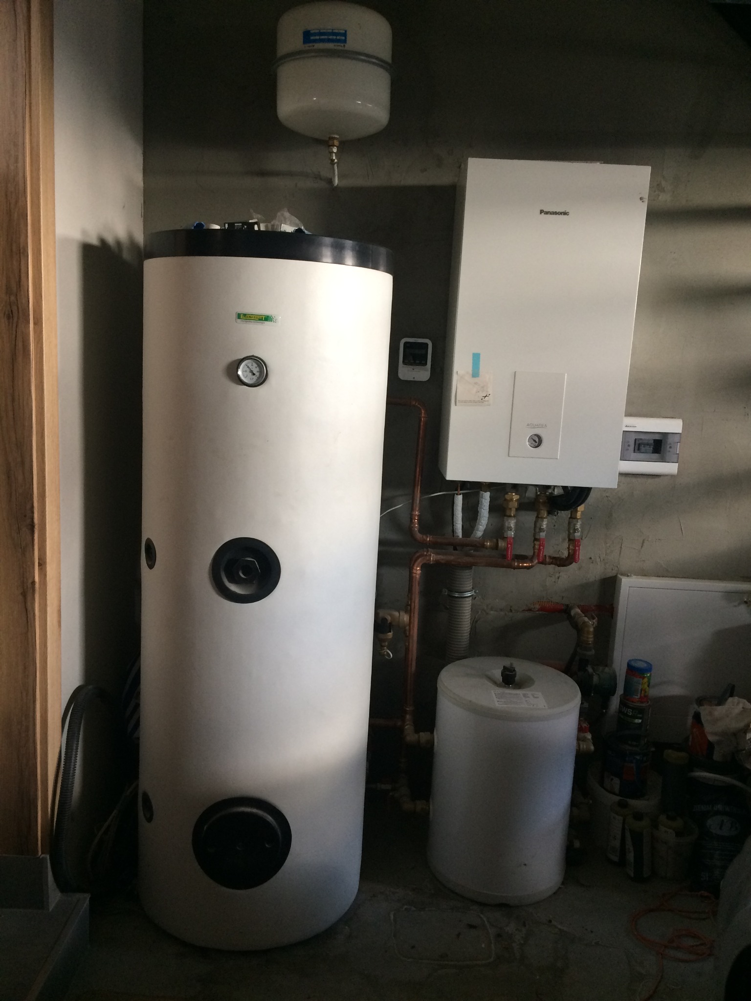 Biały zasobnik ciepłej wody użytkowej obok wiszącej jednostki wewnętrznej w kotłowni z widocznymi rurami miedzianymi i dodatkowym zbiornikiem wyrównawczym.