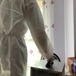 Pracownik w kombinezonie ochronnym obsługuje przemysłowy osuszacz powietrza w pomieszczeniu z zasłonami w kwiaty.