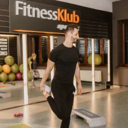 Mężczyzna w czarnym stroju rozciąga nogę w jasnym wnętrzu klubu fitness, w tle widoczne lustra, piłki do ćwiczeń i logo FitnessKlub Spa.