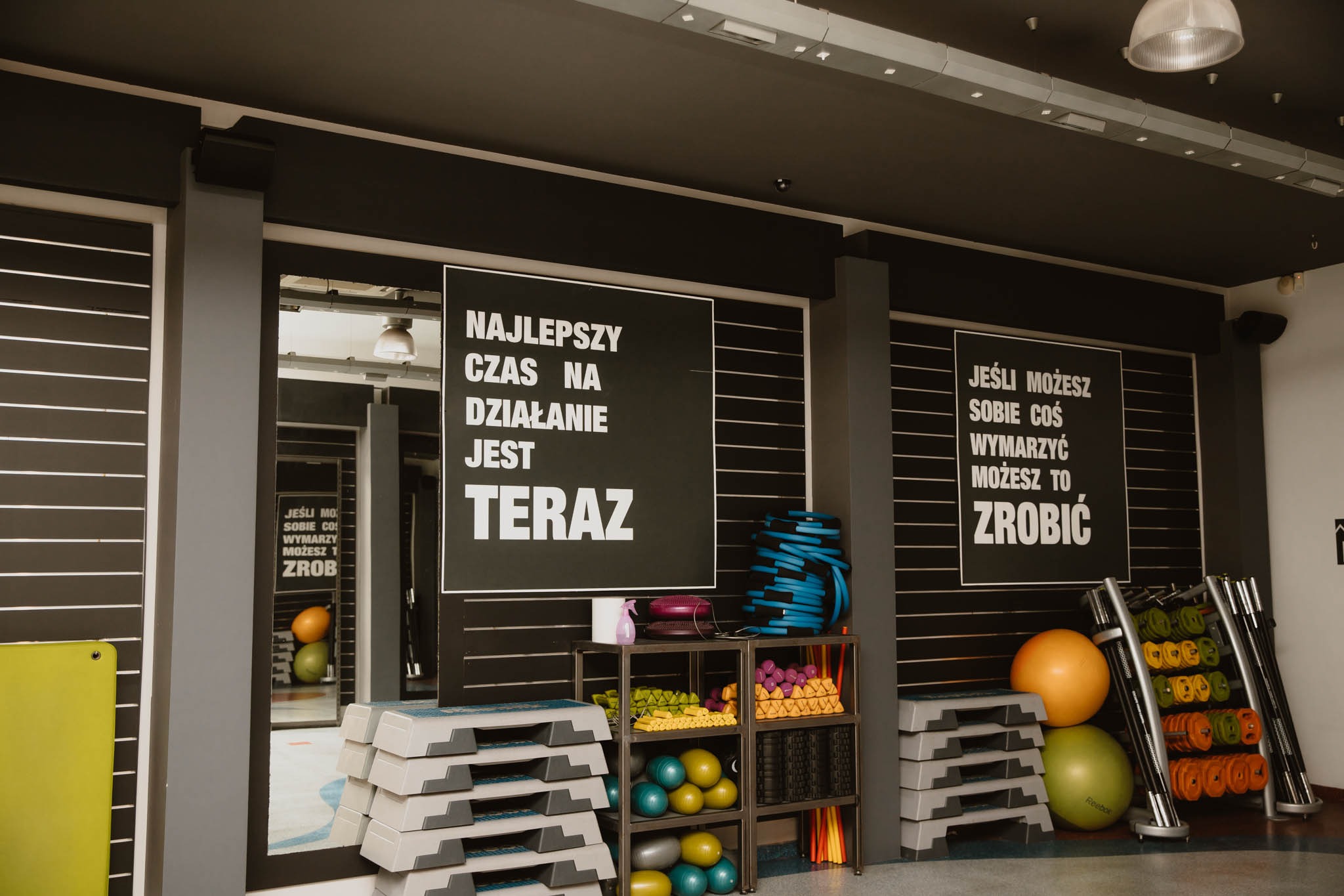 Wnętrze siłowni z motywacyjnymi hasłami na ścianach, regałami z ciężarkami, piłkami i stepami do ćwiczeń, widok z perspektywy osoby stojącej na sali.