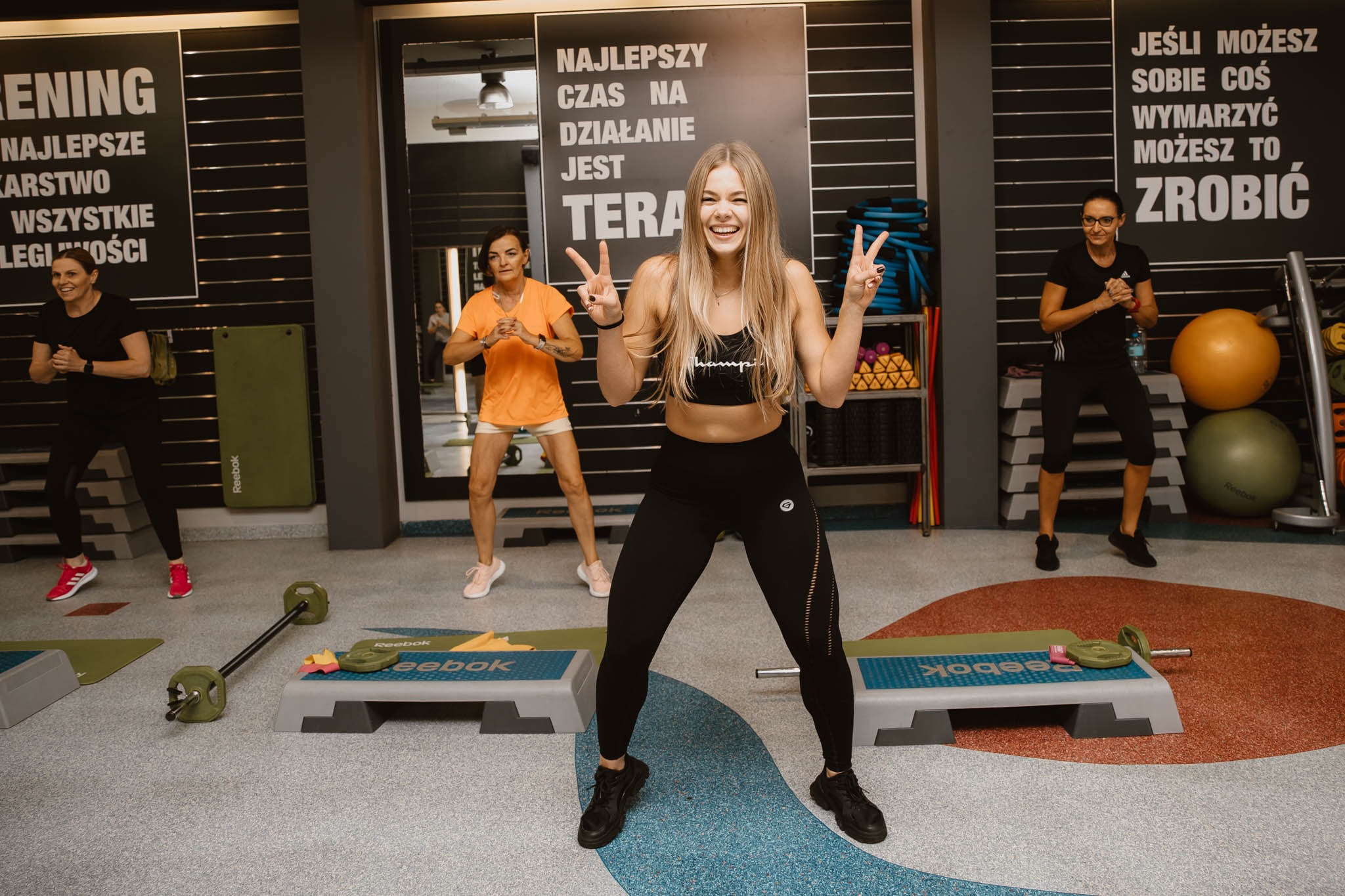 Grupa kobiet ćwiczy w siłowni, uśmiechnięta blondynka w czarnym stroju sportowym na pierwszym planie, pokazująca gest zwycięstwa, w tle widoczne stepy, ciężary i motywacyjny napis na ścianie.
