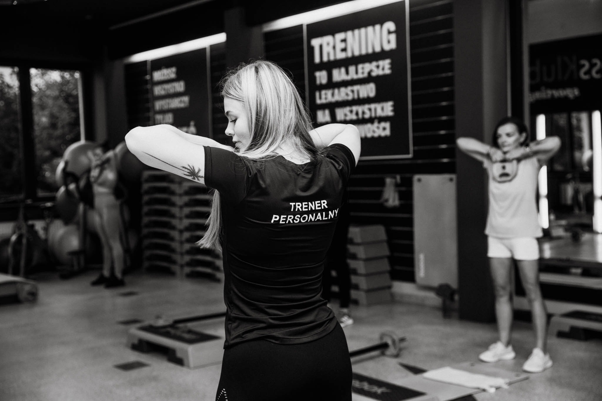 Kobieta w czarnej koszulce z napisem 'TRENER PERSONALNY' na plecach, ćwicząca w siłowni w Lesznie. Widoczny tatuaż palmy na ramieniu, w tle druga osoba ćwicząca z kettlebellem.