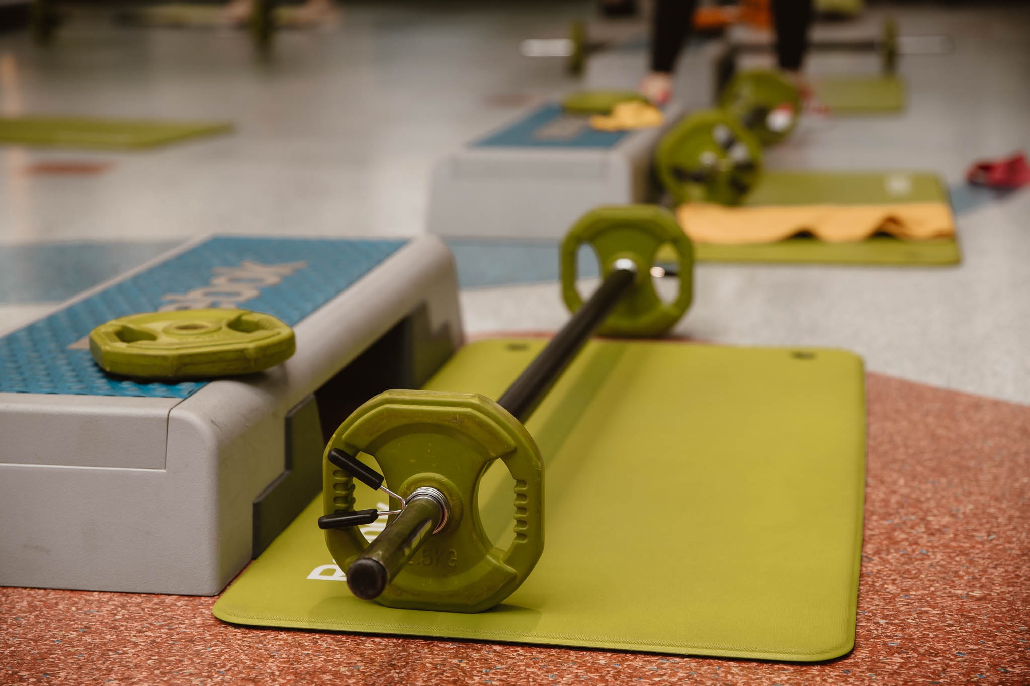 Sala fitness z zielonymi obciążeniami i stepem, na podłodze leżą maty do ćwiczeń, w tle widoczne osoby ćwiczące.