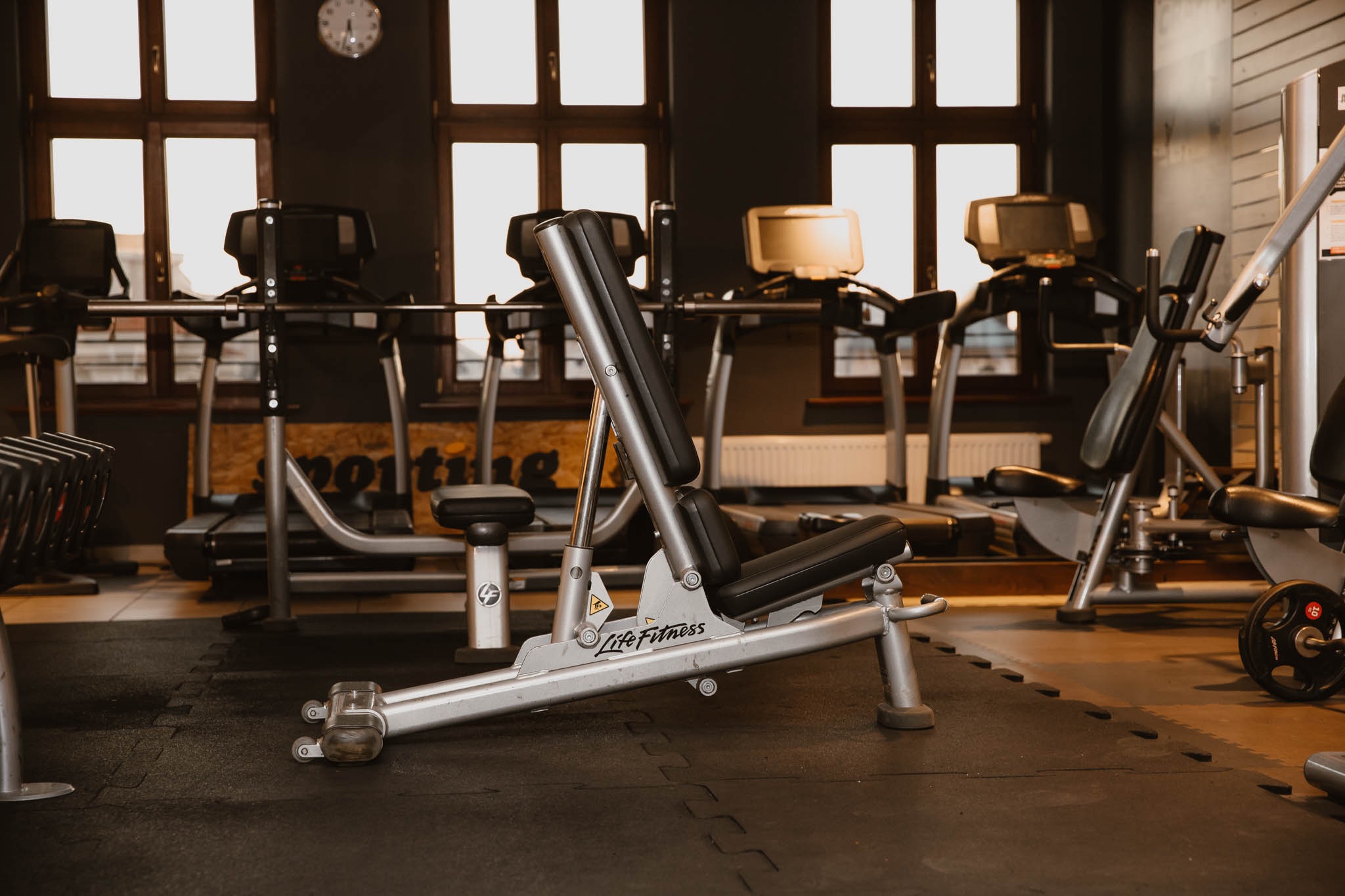 Wnętrze siłowni z widocznym sprzętem do ćwiczeń: ławka do wyciskania, bieżnie i maszyny siłowe. Na ścianie w tle okna i zegar.