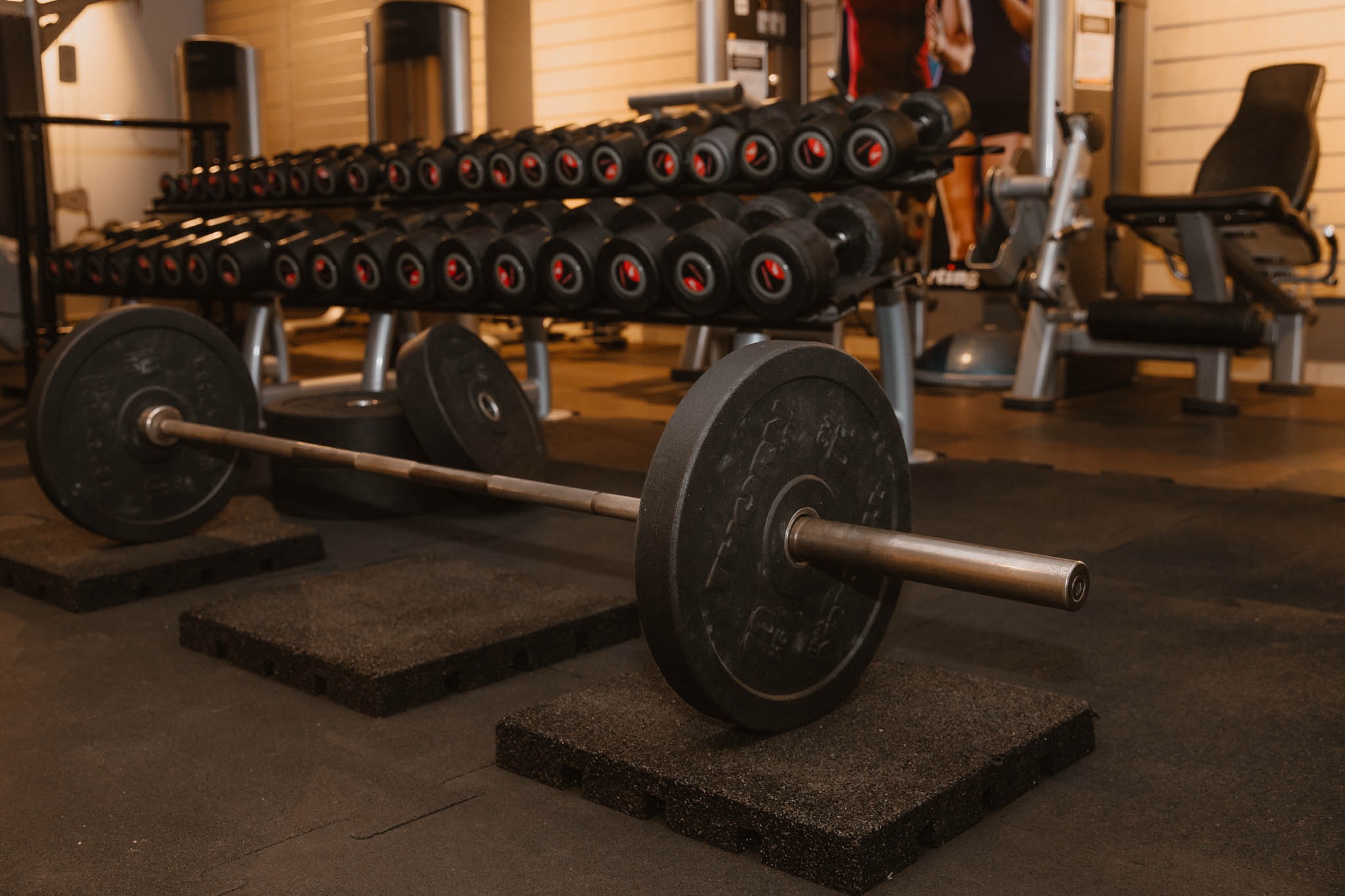 Sztanga z obciążeniem na czarnych matach w siłowni, w tle regał z hantlami i maszyna do ćwiczeń.
