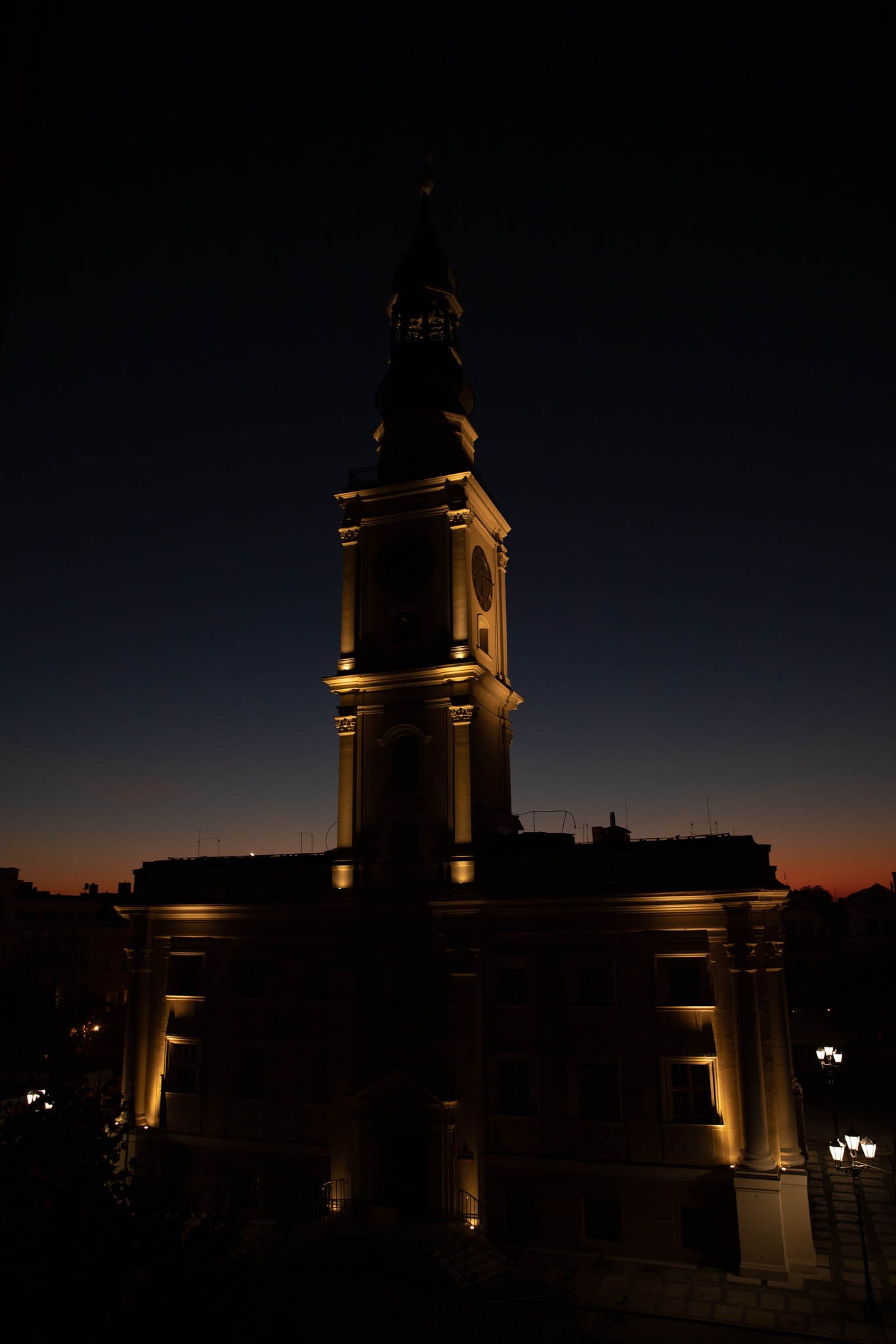 Oświetlony budynek z wieżą zegarową na tle ciemniejącego nieba o zmierzchu, widoczne detale architektoniczne i lampy uliczne.