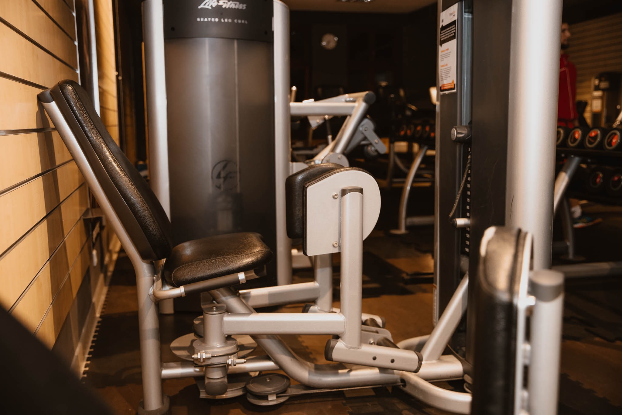 Siłownia: maszyna do ćwiczeń na nogi z czarnym siedziskiem i srebrną ramą, w tle inne urządzenia i ciężary.
